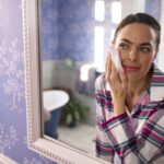 Woman At Home Wearing Pyjamas Looking In Bathroom Mirror Taking Off Make Up With Wipe